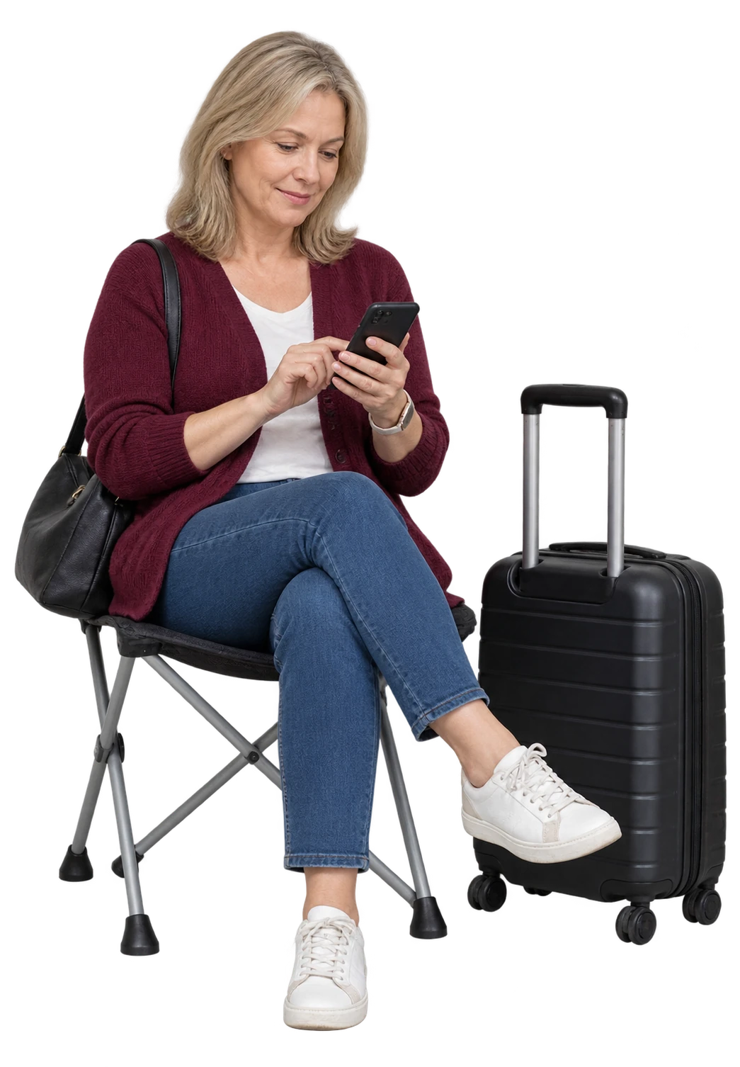 Woman in a burgundy cardigan checking her phone on a folding chair beside a black suitcase