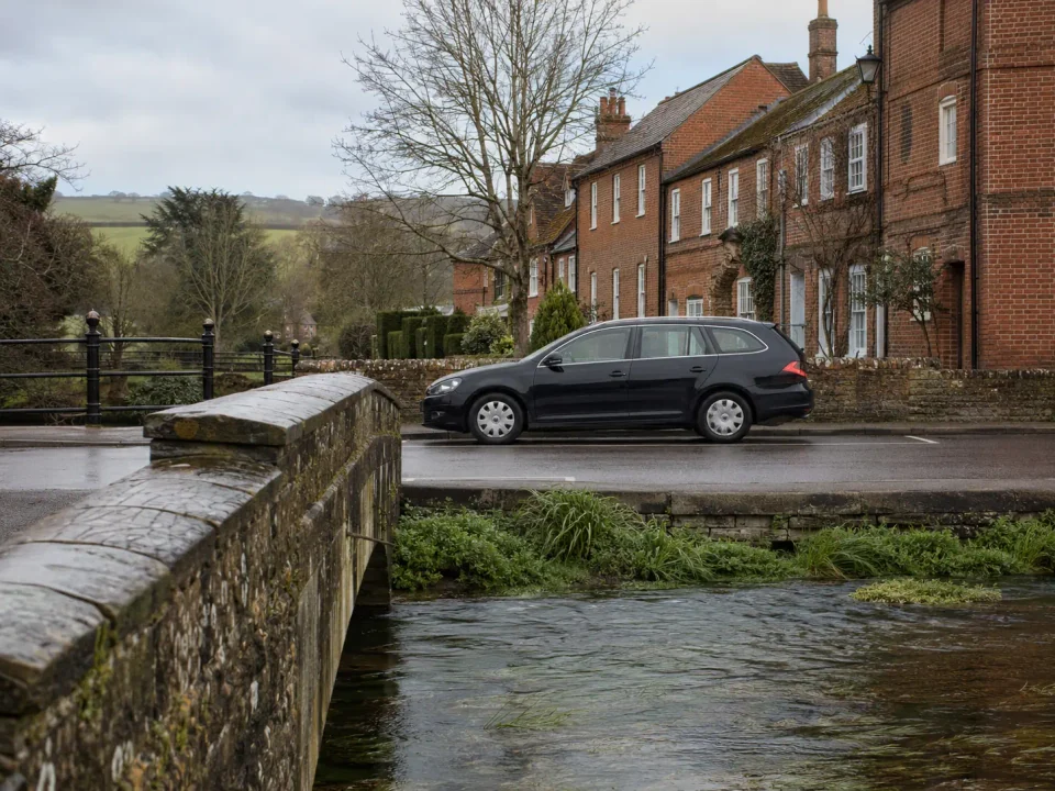 Airport transfer vehicle parked beside a chalk stream and red-brick Andover street