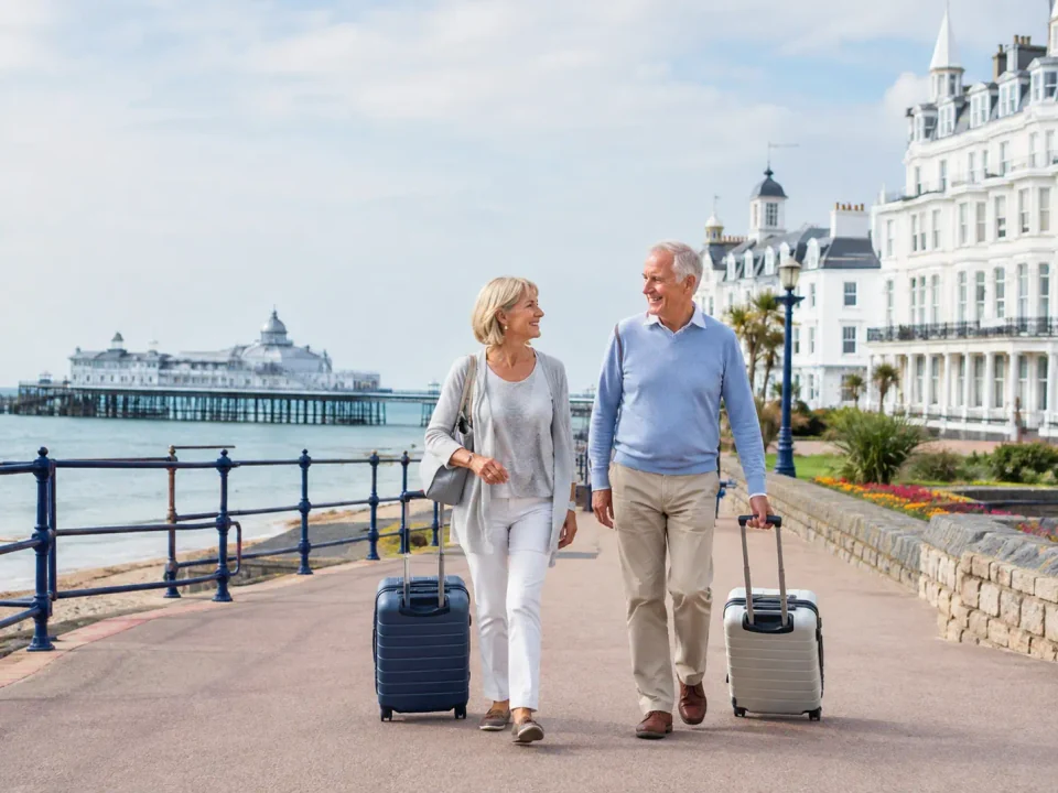 Older travelling couple with suitcases on Eastbourne seafront beside the pier