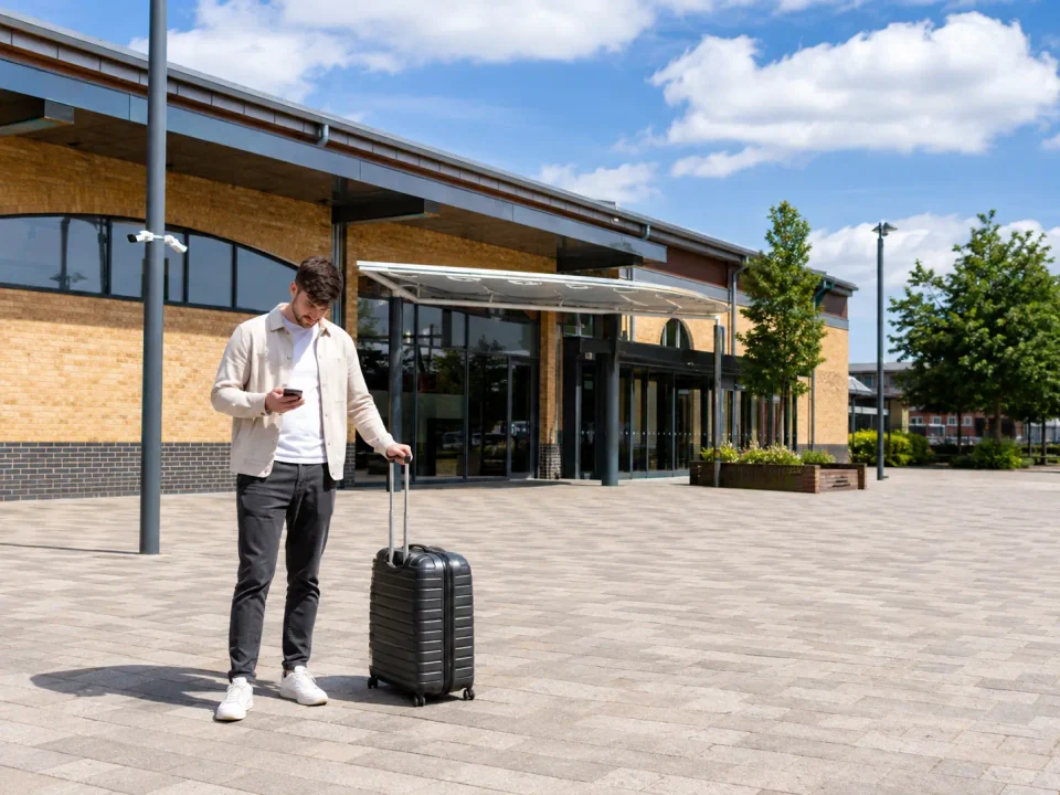 Solo traveller checking a phone with a suitcase outside a modern Feltham station