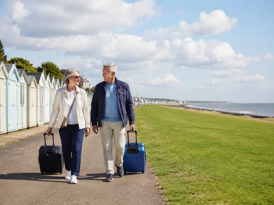 Older travelling couple with suitcases beside the greensward and beach huts in Frinton-on-Sea
