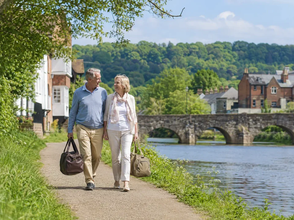 Older travellers with weekend bags beside the River Mole and historic bridge in Leatherhead