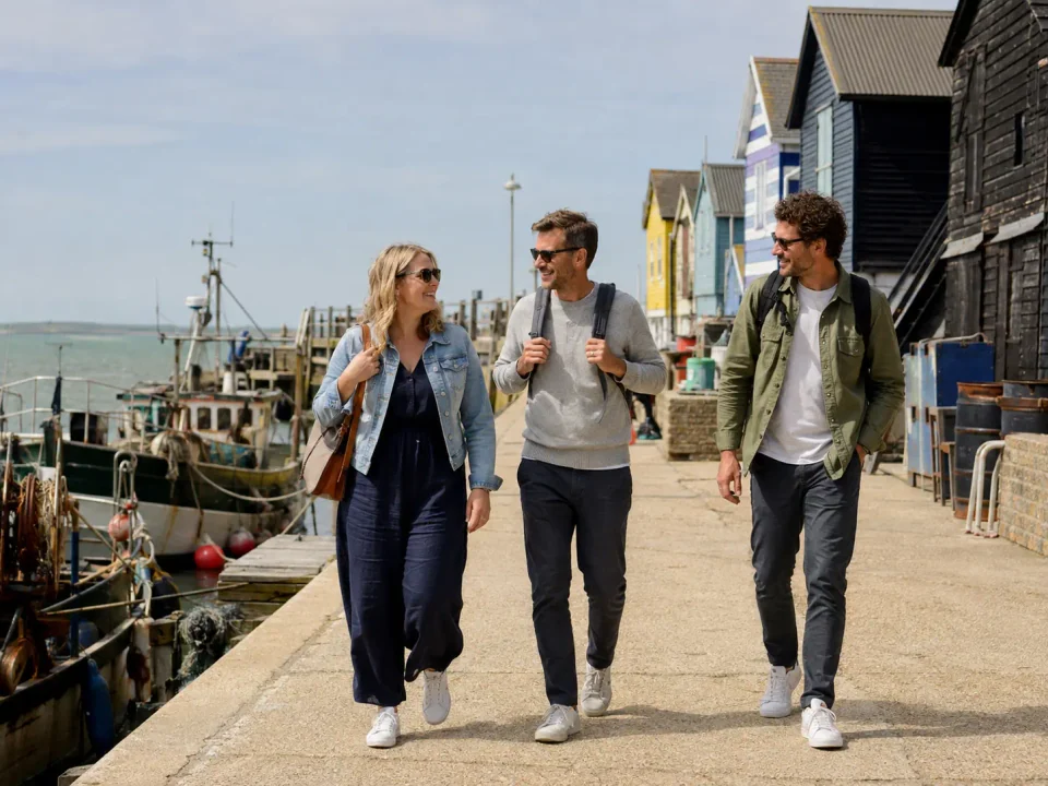 Friends walking beside fishing boats and colourful harbour huts in Whitstable